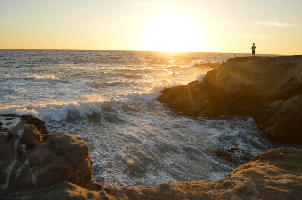 A man stands on a cliff above a bay as the sun sets.