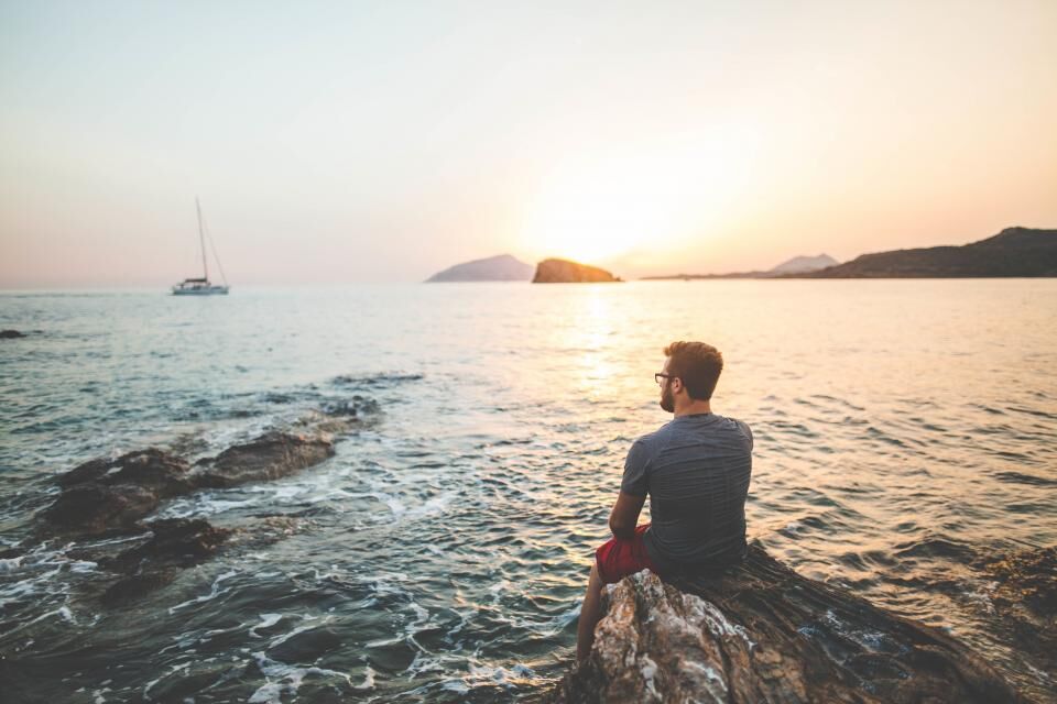 A man sits alone on the rocks at the edge of the sea at sunset.