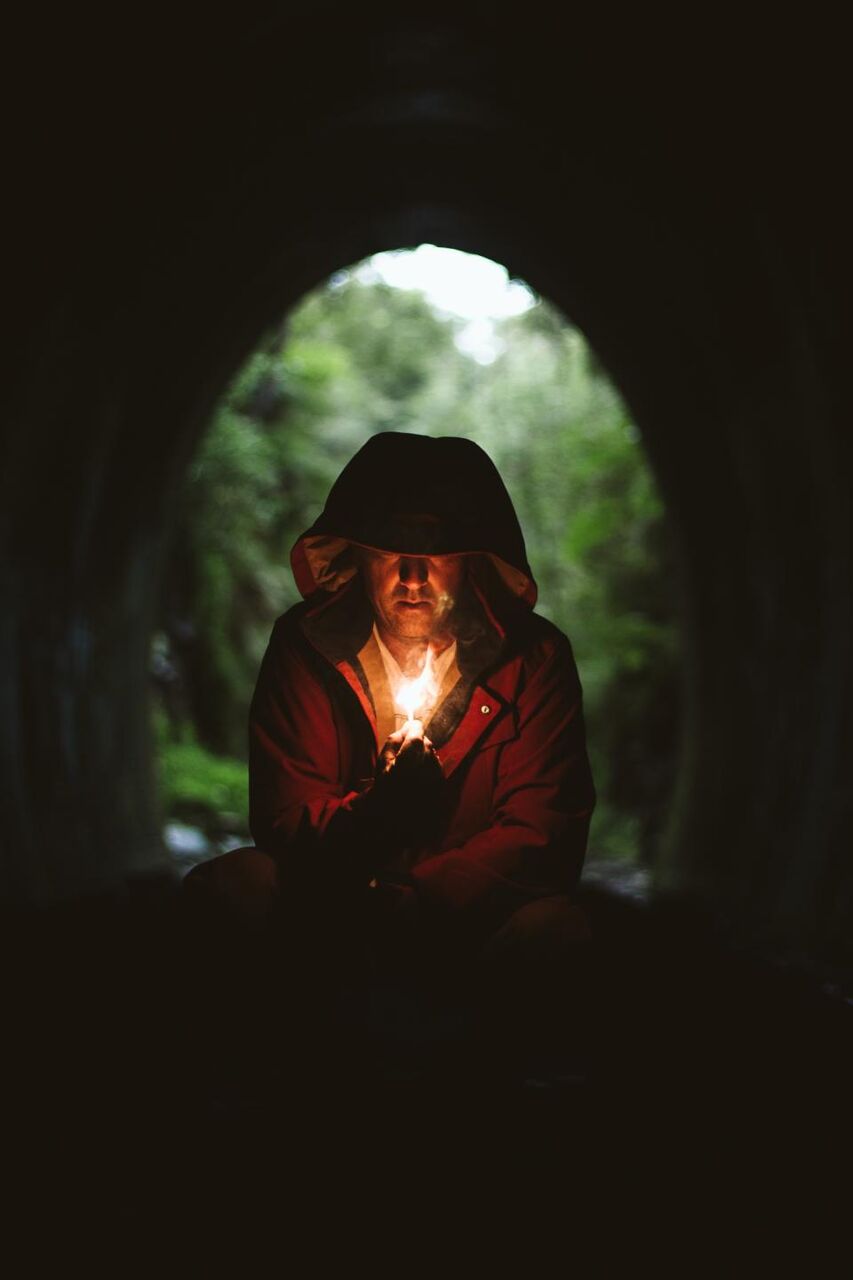 A man in a hood holds a small light in the dark.