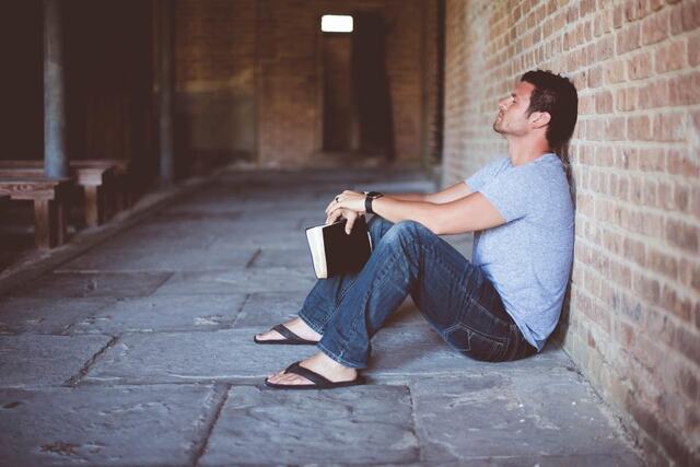 A man sits on the ground against a brick wall, looking up.