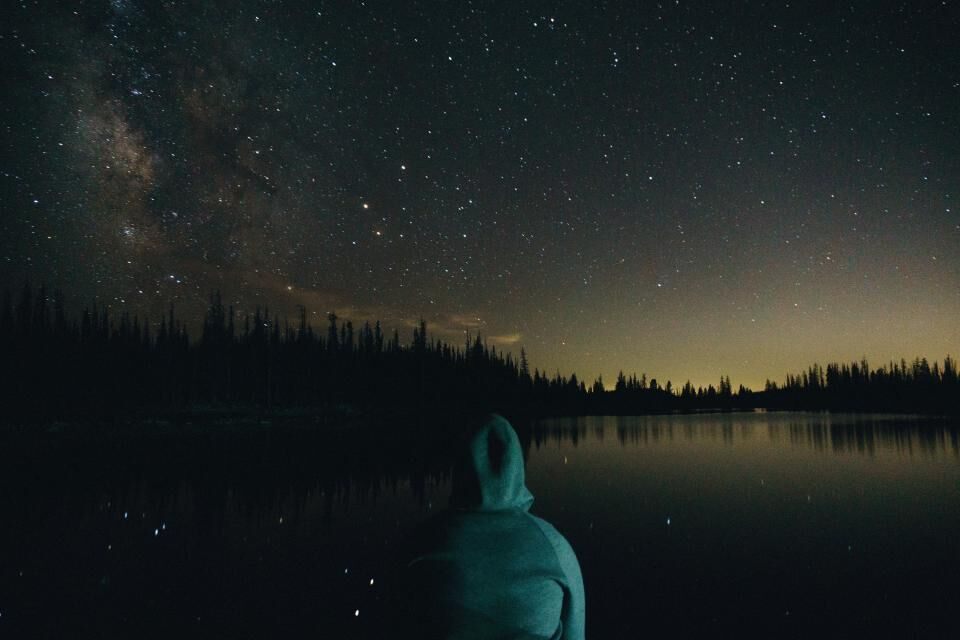 A person in a hood sits by a lake under a night sky.