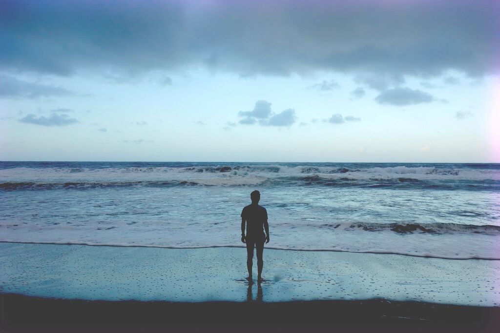The silhouette of a man standing in shallow water, facing the horizon.