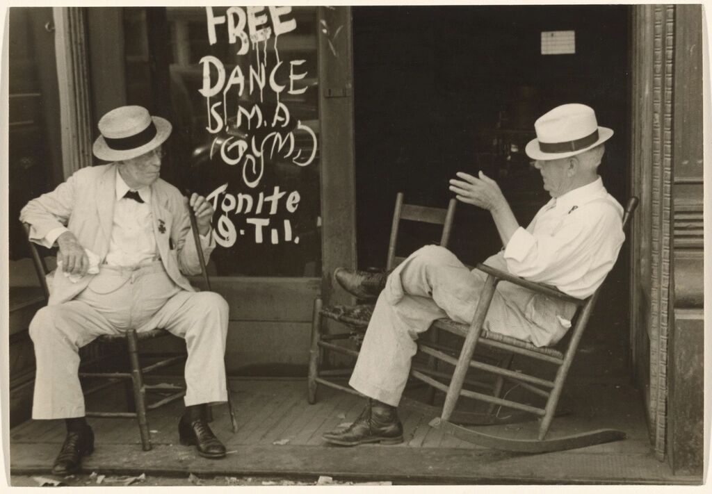 Two older men sit and talk outside a doorway.